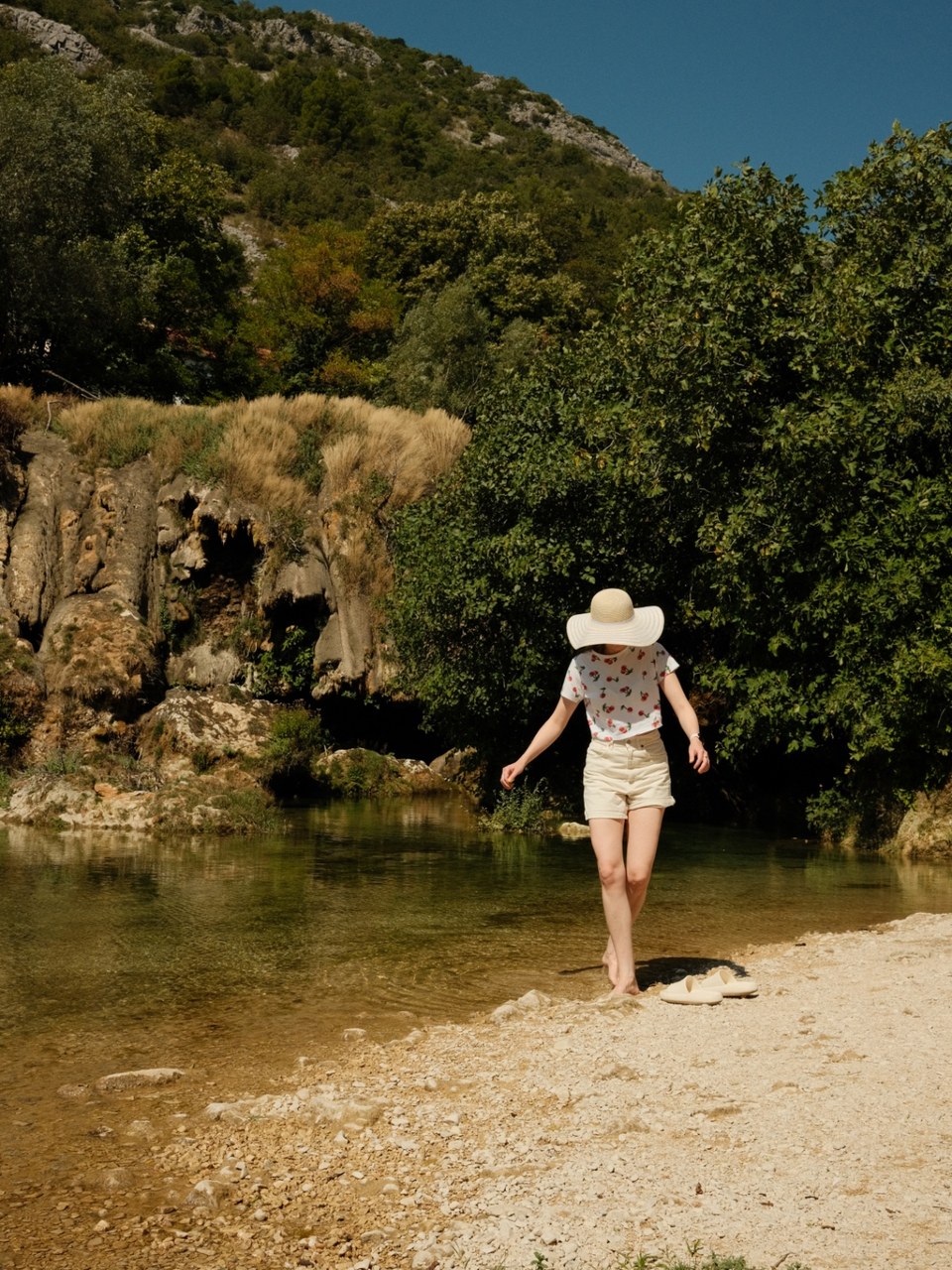 Woman in a wide-brimmed hat wading barefoot along a shallow riverbank
