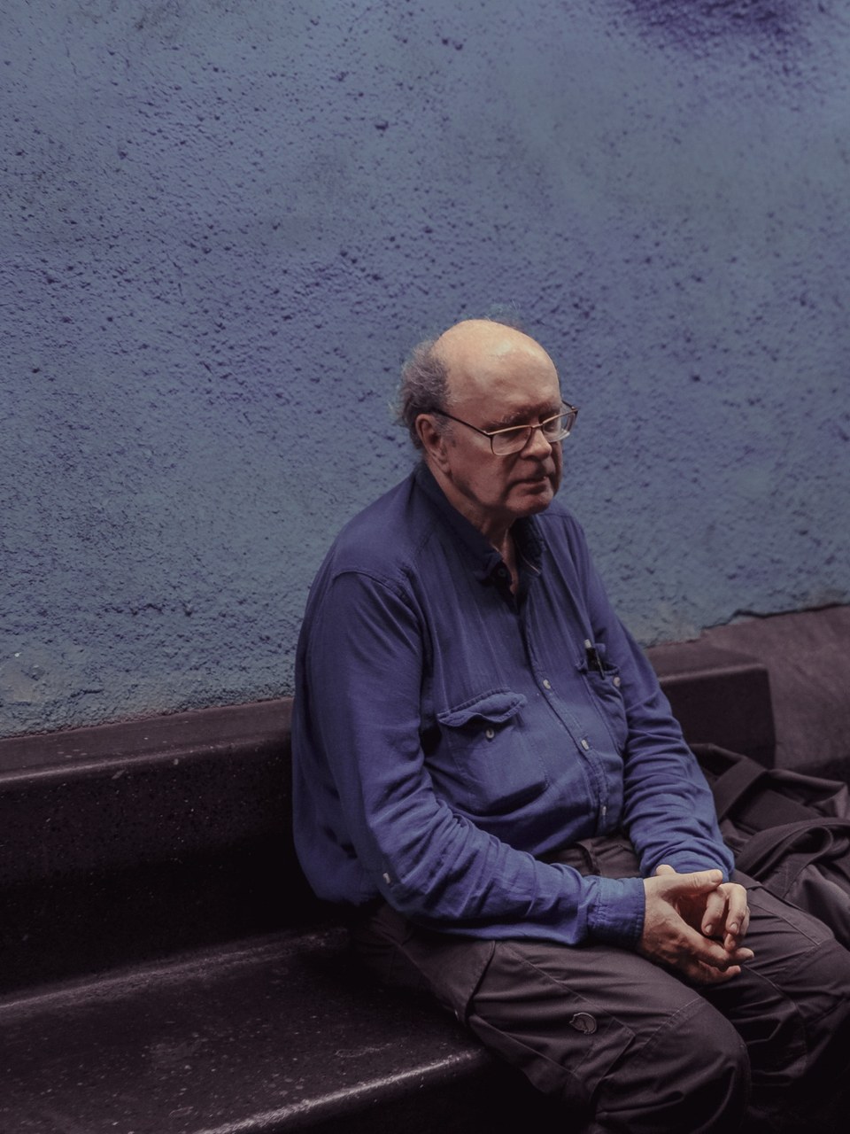 Older man in glasses seated with clasped hands against a blue wall