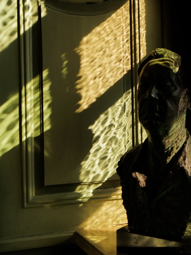 Bronze bust on a mantel with rippled sunlight across a sage paneled wall