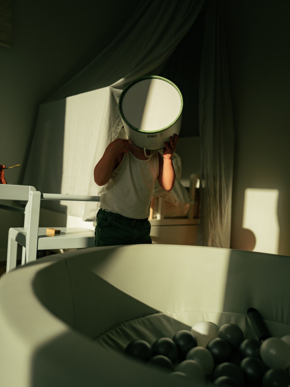 Toddler with a box over their head beside a ball pit in soft window light