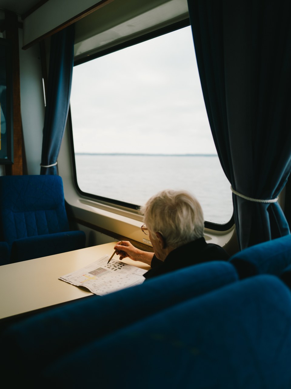 Older man doing a crossword at a table beside the sea view on a ferry