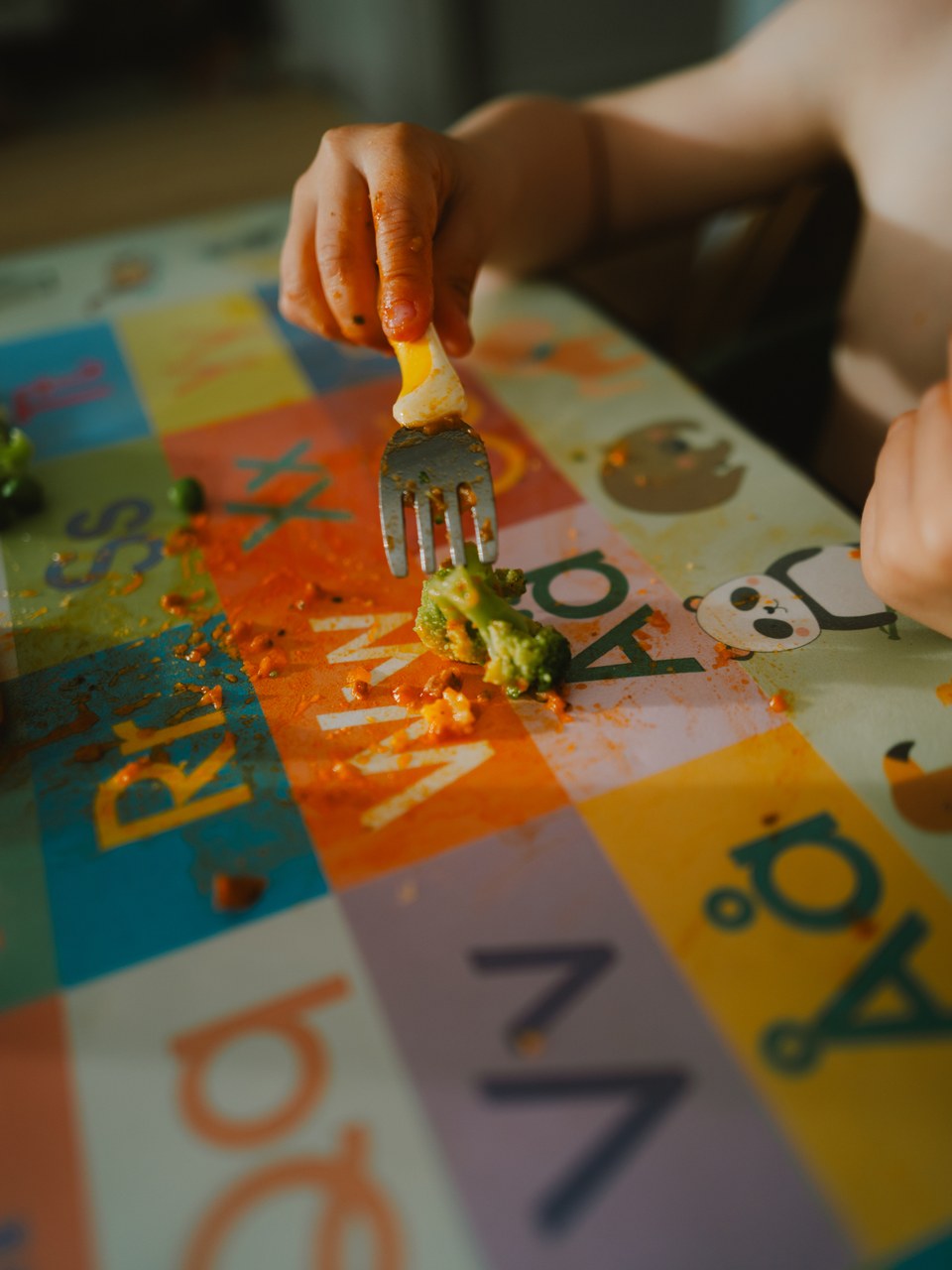 Child's hand spearing broccoli on a colourful alphabet placemat