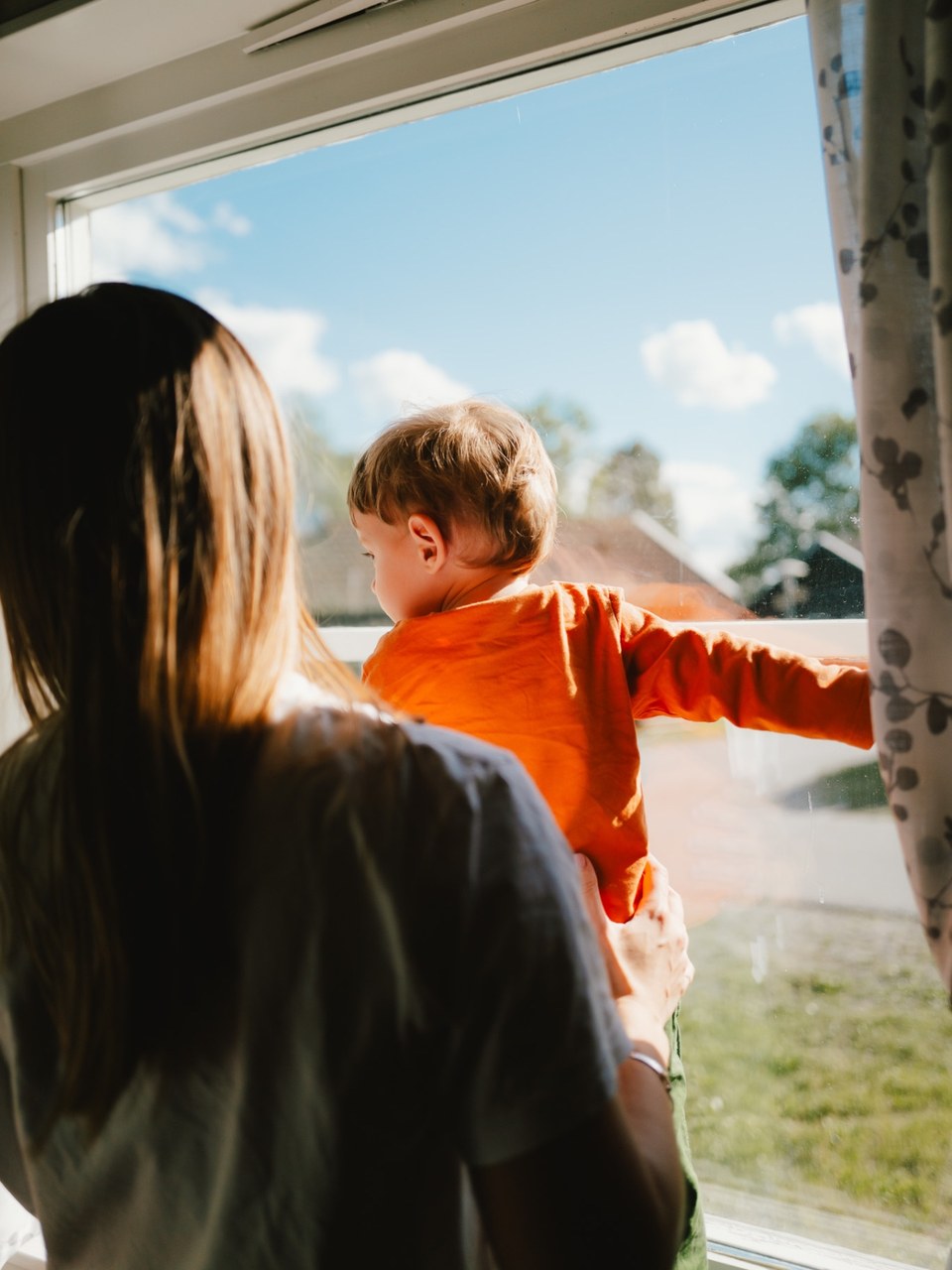 Mother holding a child in an orange shirt up to a sunlit window